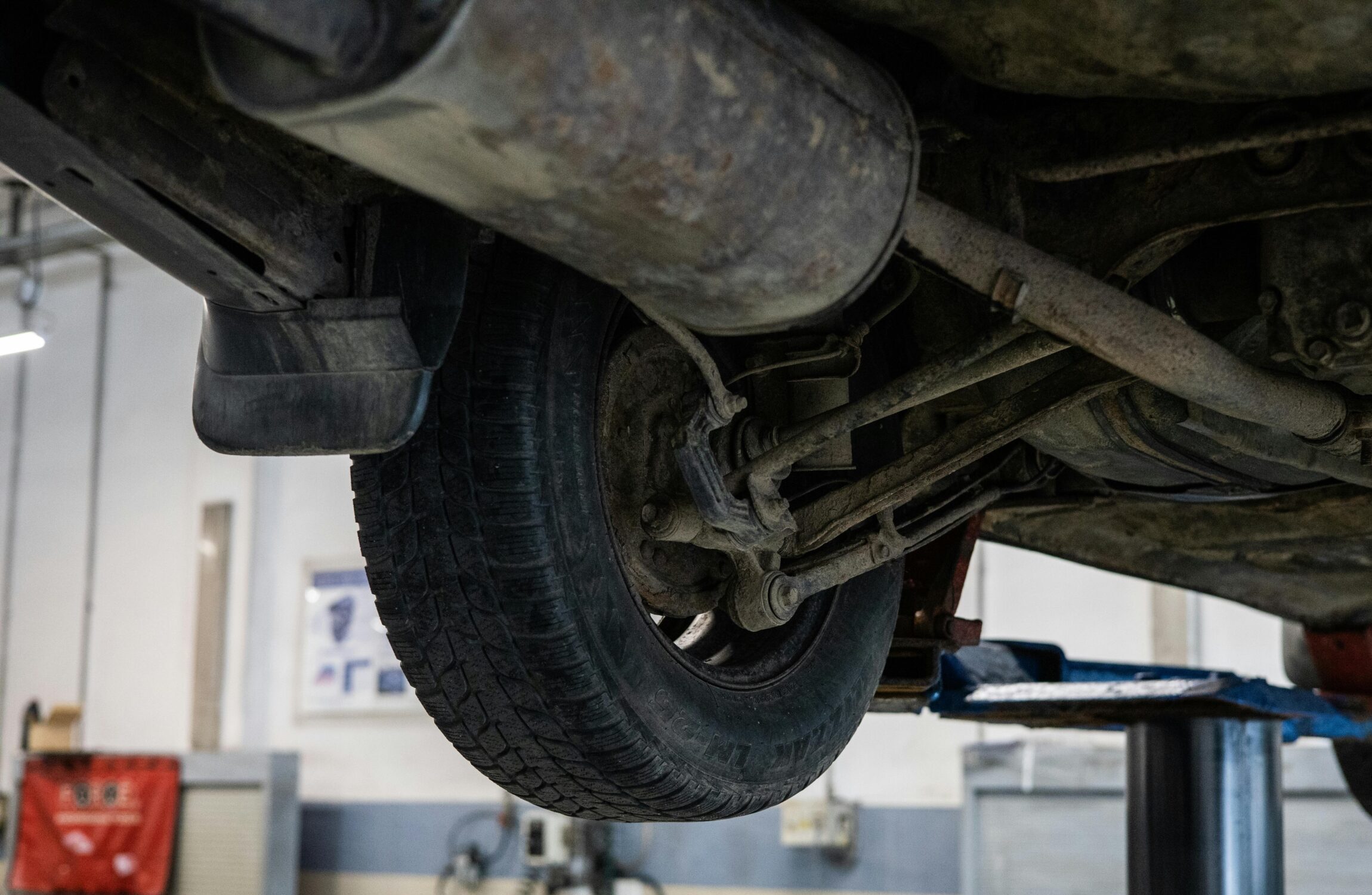 Widok podwozia auta uniesionego na podnośniku – widoczna tylna oś, zawieszenie, tłumik i koło podczas kontroli w warsztacie samochodowym.