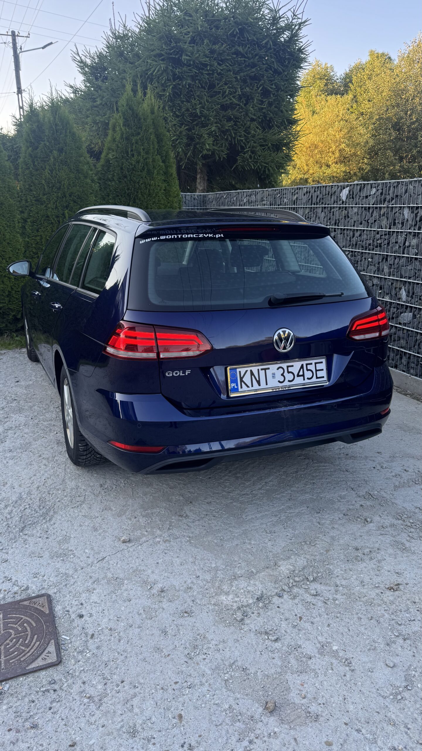 Granatowy Volkswagen Golf kombi stoi tyłem na jasnym, żwirowo-betonowym podłożu, obok wysokich zielonych tui i ogrodzenia z gabionów, w słoneczny dzień.