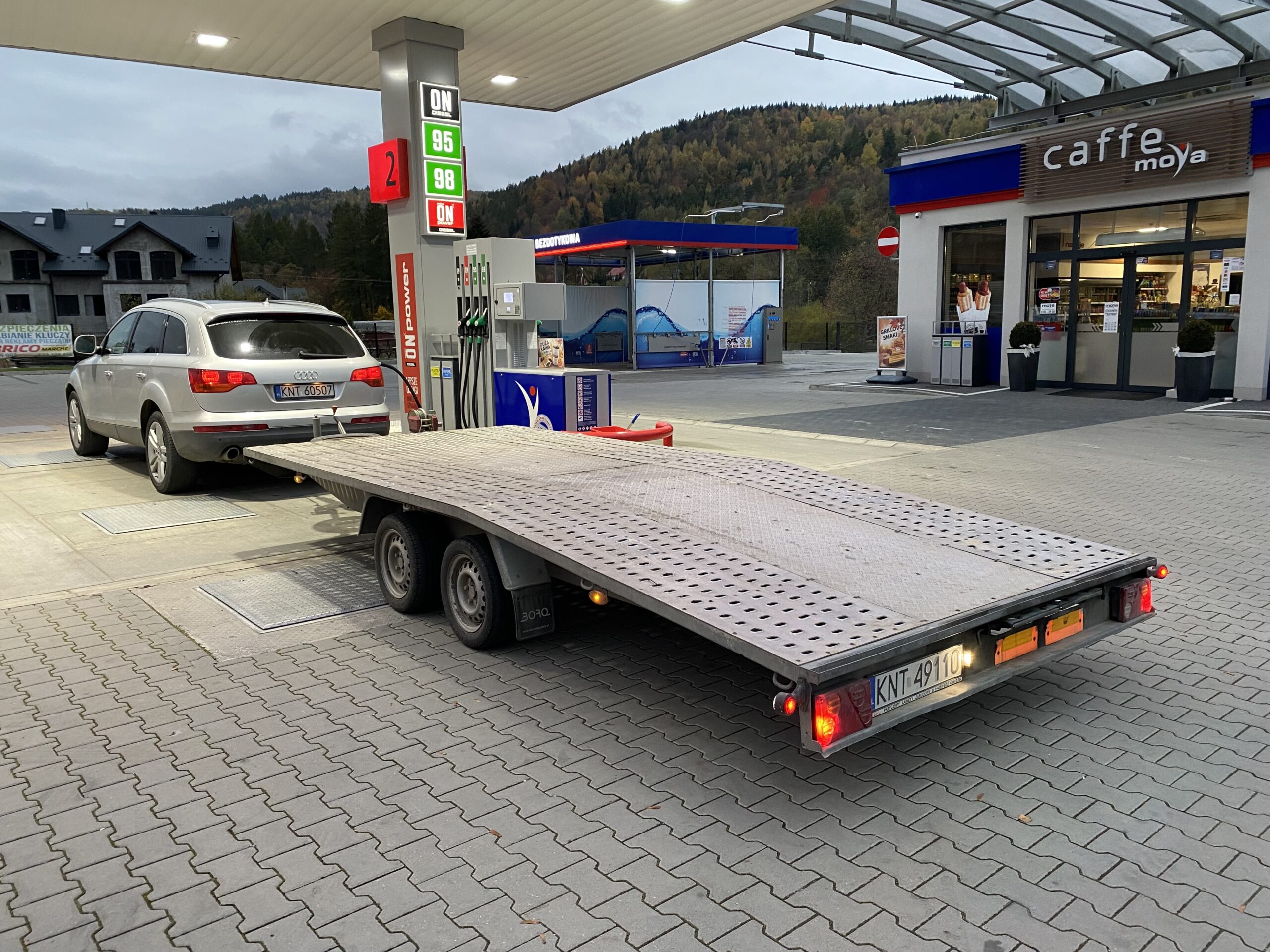 Laweta z podpiętym srebrnym SUV-em stoi przy dystrybutorach na stacji paliw; w tle widać budynek sklepu, oznaczenia ceny paliw oraz górskie zbocza pokryte jesienną roślinnością.