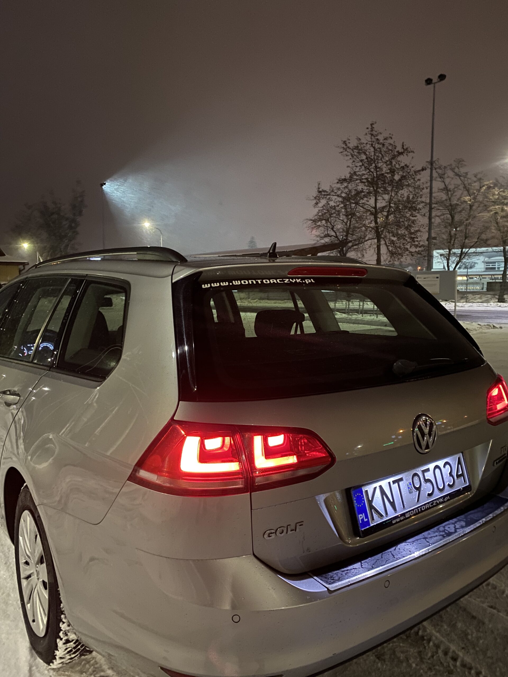 Srebrny Volkswagen Golf z włączonymi tylnymi światłami stoi na ośnieżonym parkingu nocą; w tle widać drzewa, latarnie oraz padający śnieg oświetlony silnym reflektorem.