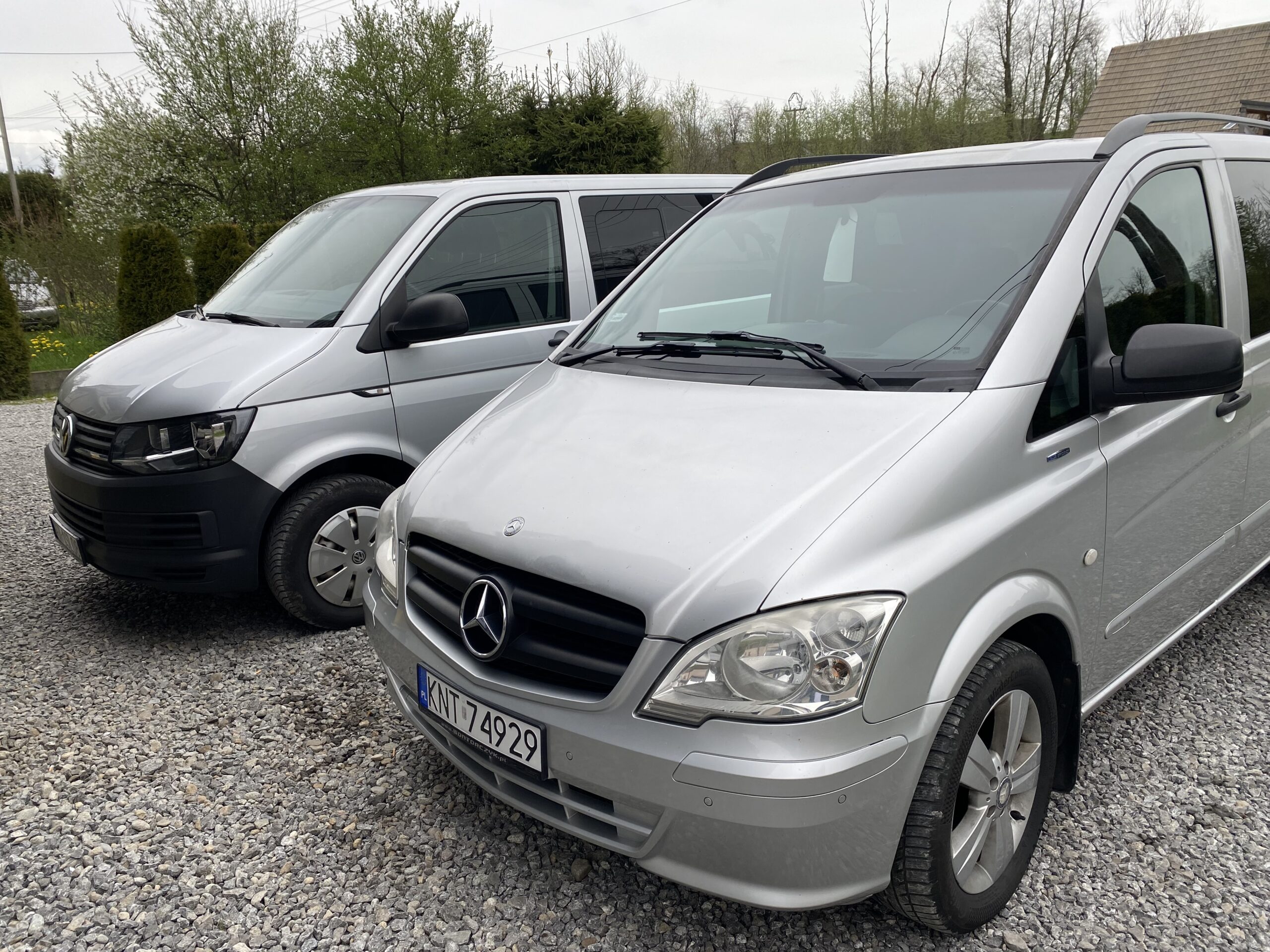 Dwa srebrne busy – Mercedes Vito z przodu oraz Volkswagen Transporter z tyłu – stoją obok siebie na żwirowym placu, w otoczeniu drzew i niskiej zabudowy.