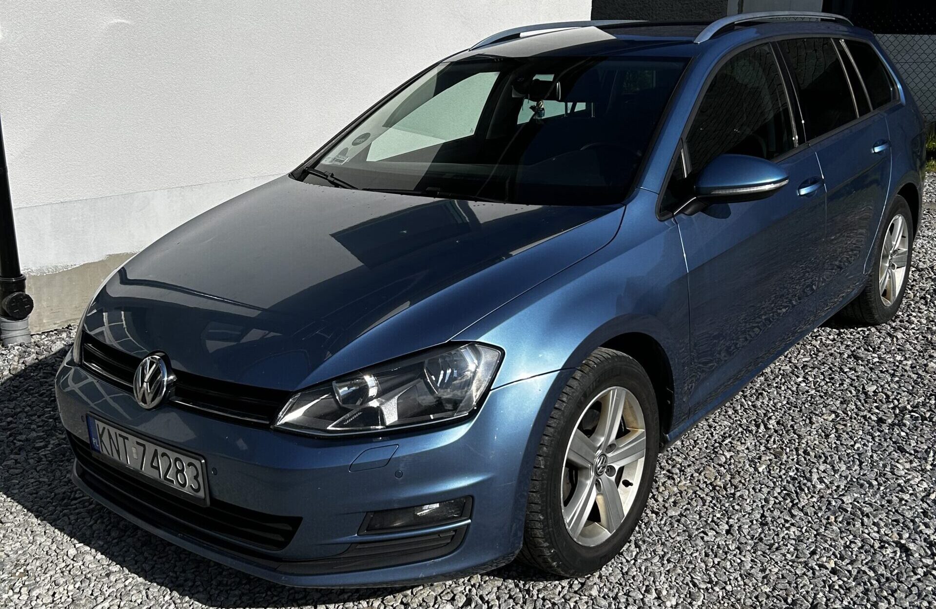 Niebieski kombi Volkswagen stoi przodem przy jasnej elewacji budynku, zaparkowany na żwirowym podłożu w słoneczny dzień.