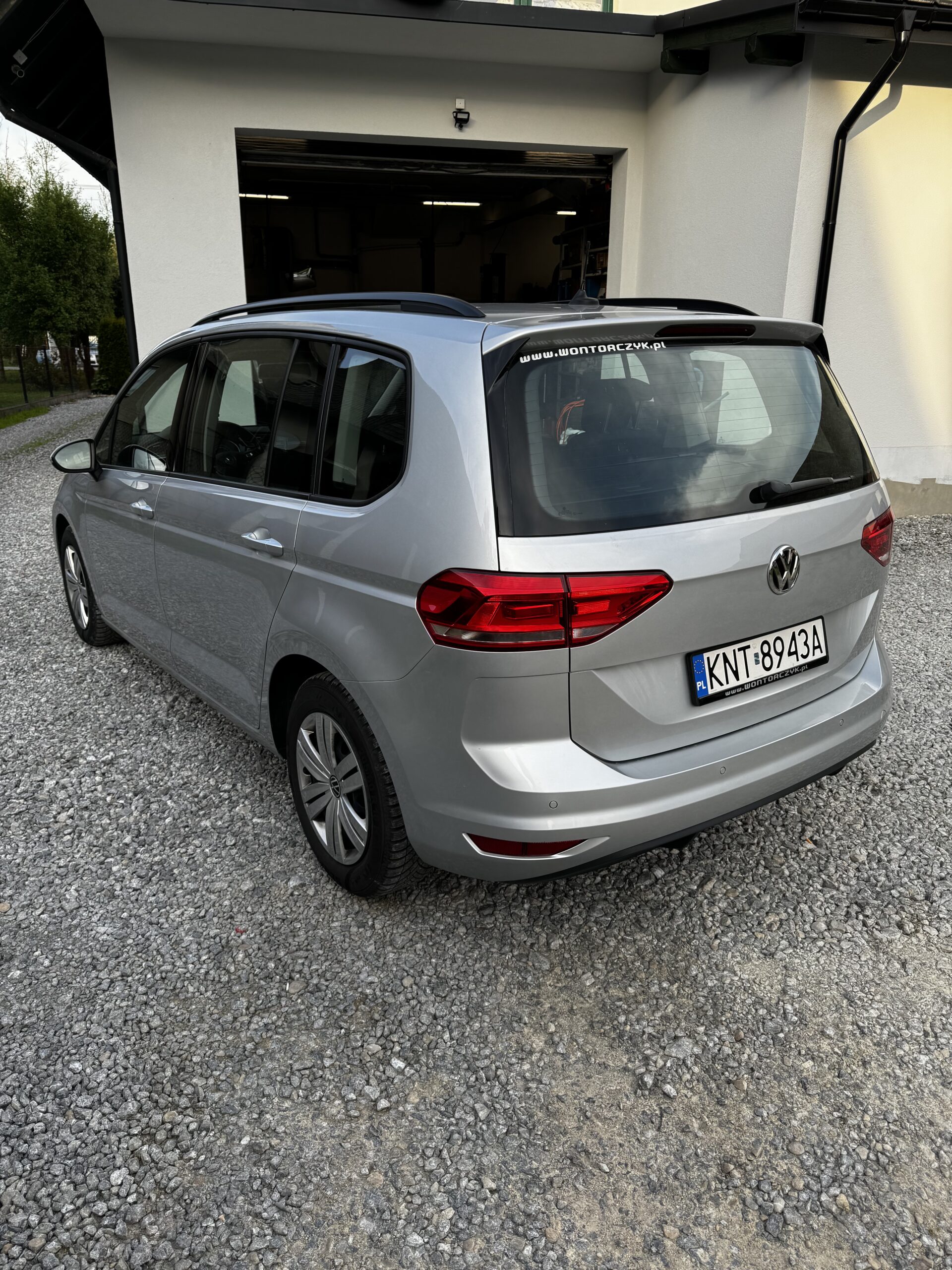 Srebrny minivan Volkswagen stoi tyłem na żwirowym podjeździe przed jasnym budynkiem z otwartym garażem.