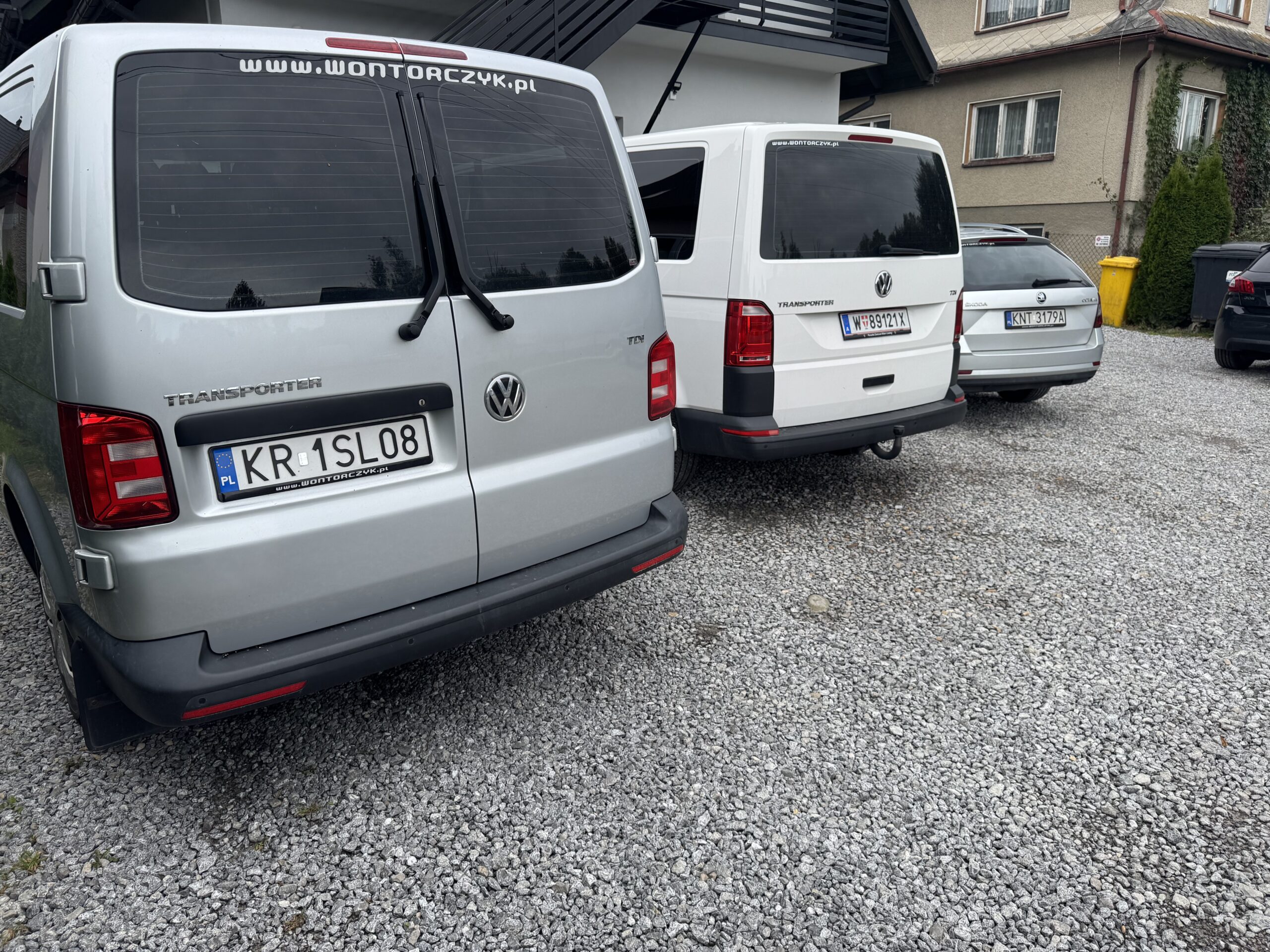 Kilka busów Volkswagen Transporter oraz samochód osobowy zaparkowane tyłem na żwirowym podjeździe przed budynkiem mieszkalnym.