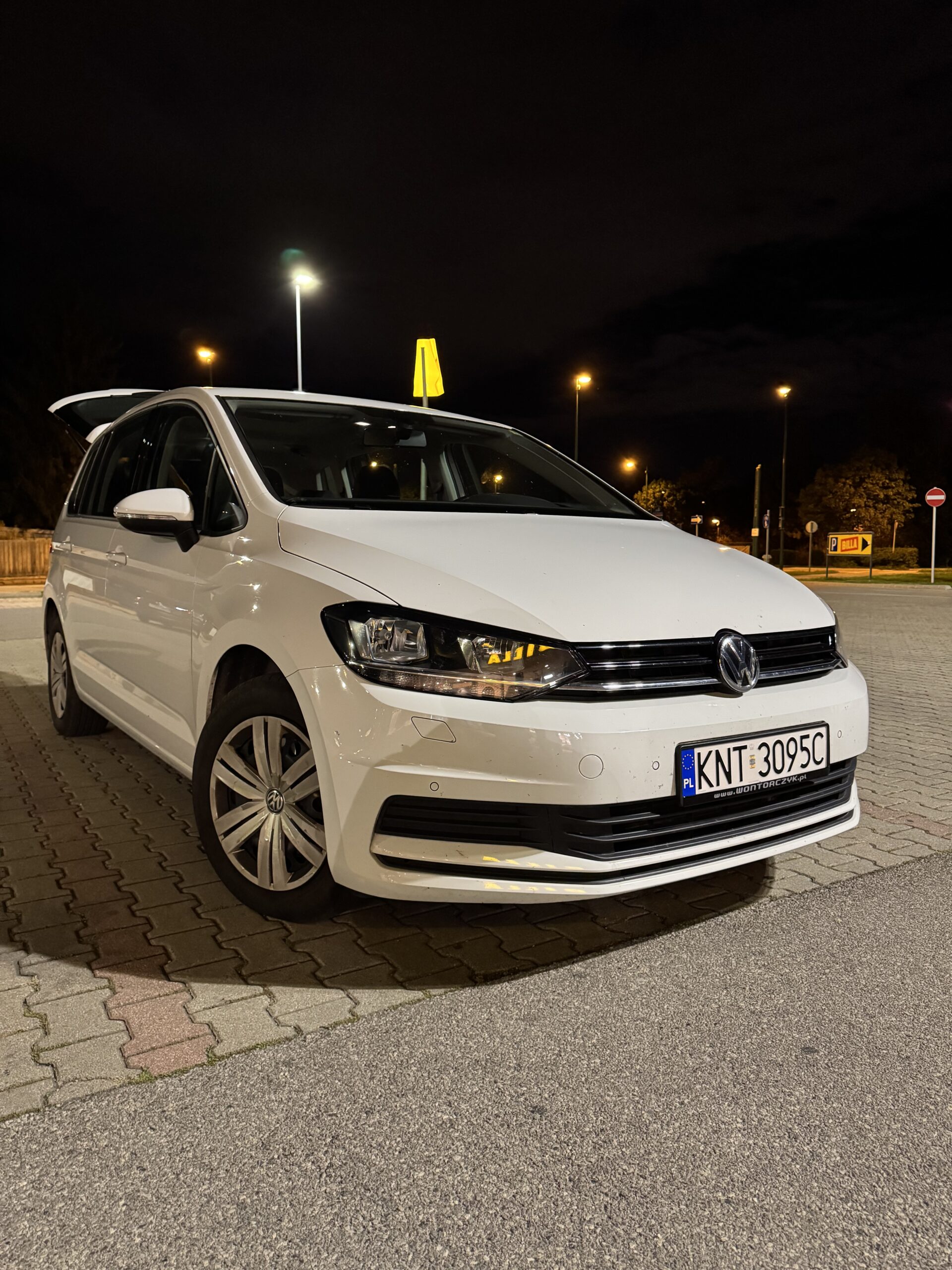 Biały Volkswagen zaparkowany na nocnym, dobrze oświetlonym parkingu, z włączonymi światłami i otwartą tylną klapą, stojący na kostce brukowej obok asfaltowej nawierzchni.