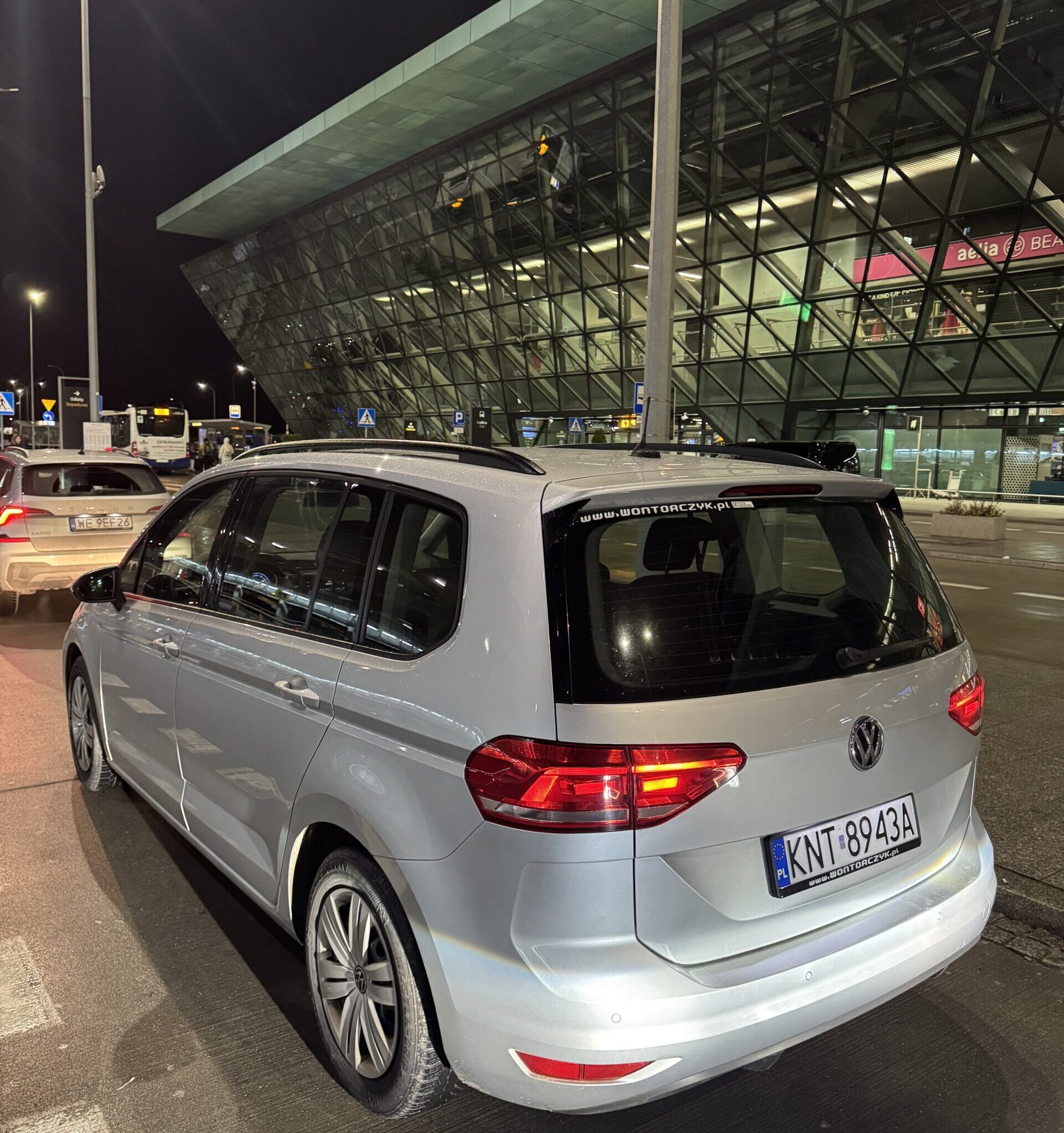 Srebrny Volkswagen Touran zaparkowany nocą przed nowoczesnym, przeszklonym terminalem lotniska, z włączonymi światłami tylnymi.