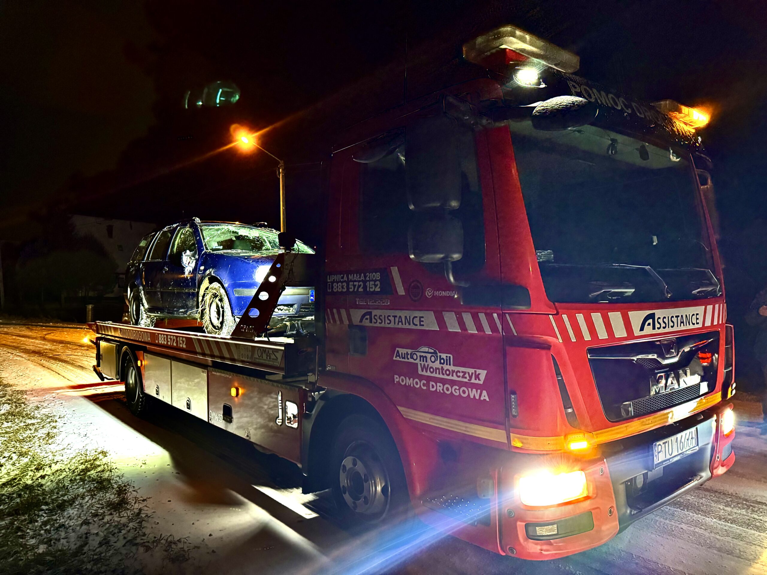 Czerwona laweta pomocy drogowej MAN stoi nocą na ośnieżonej drodze, przewożąc niebieski, uszkodzony samochód; scena oświetlona jest światłami lawety i uliczną lampą.