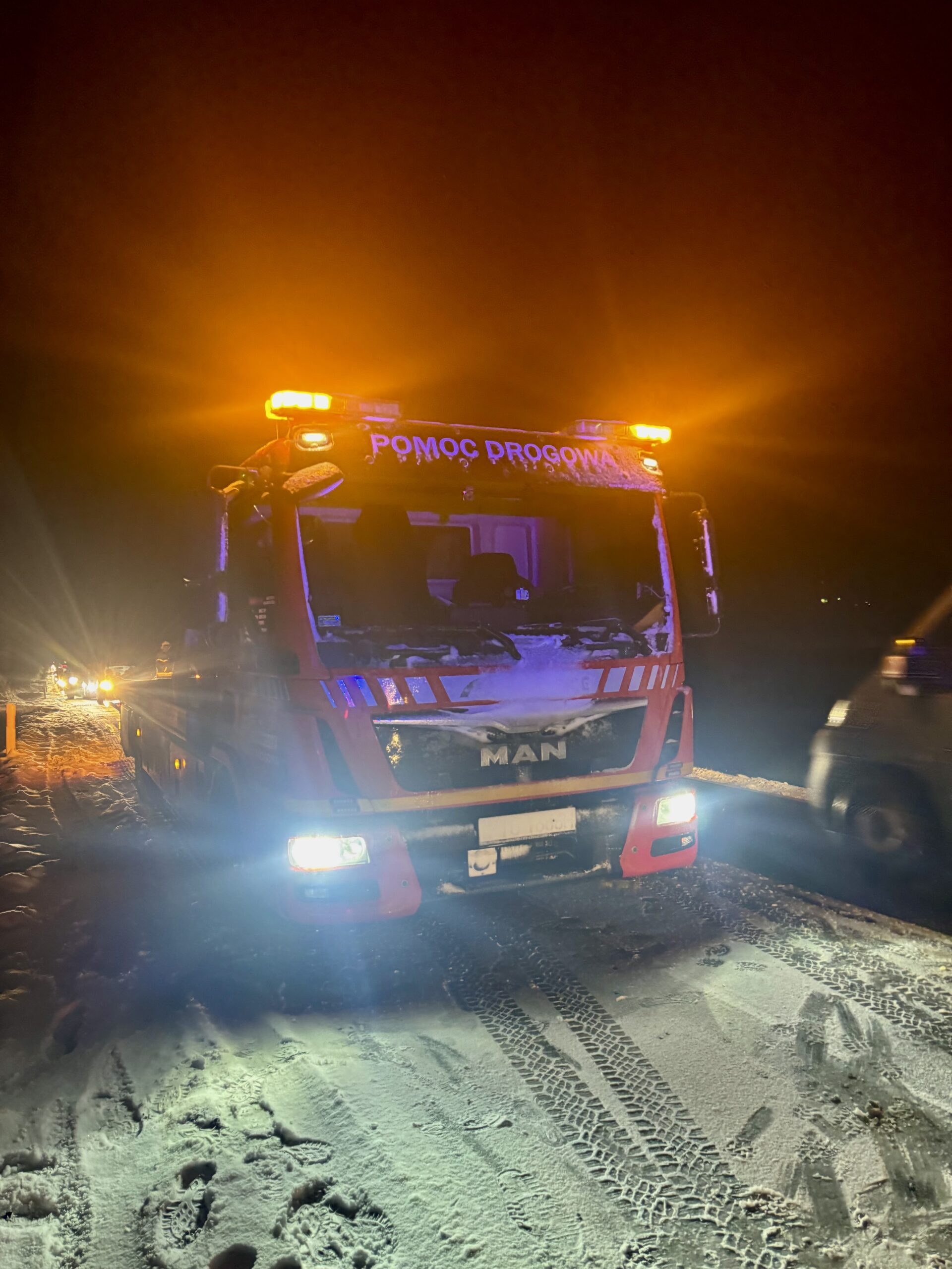 Laweta pomocy drogowej marki MAN stoi na ośnieżonej drodze nocą, z włączonymi pomarańczowymi światłami ostrzegawczymi i intensywnym oświetleniem przednim.