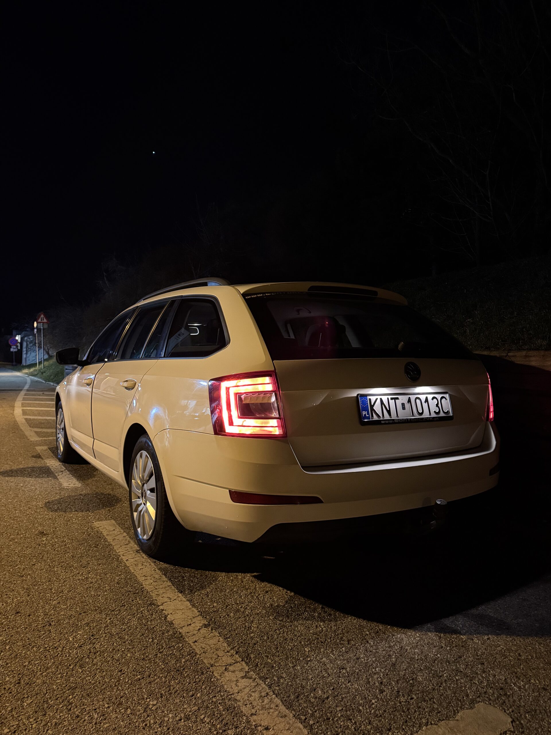 Jasna Skoda kombi zaparkowana nocą na poboczu oświetlonej drogi, z włączonymi tylnymi światłami.