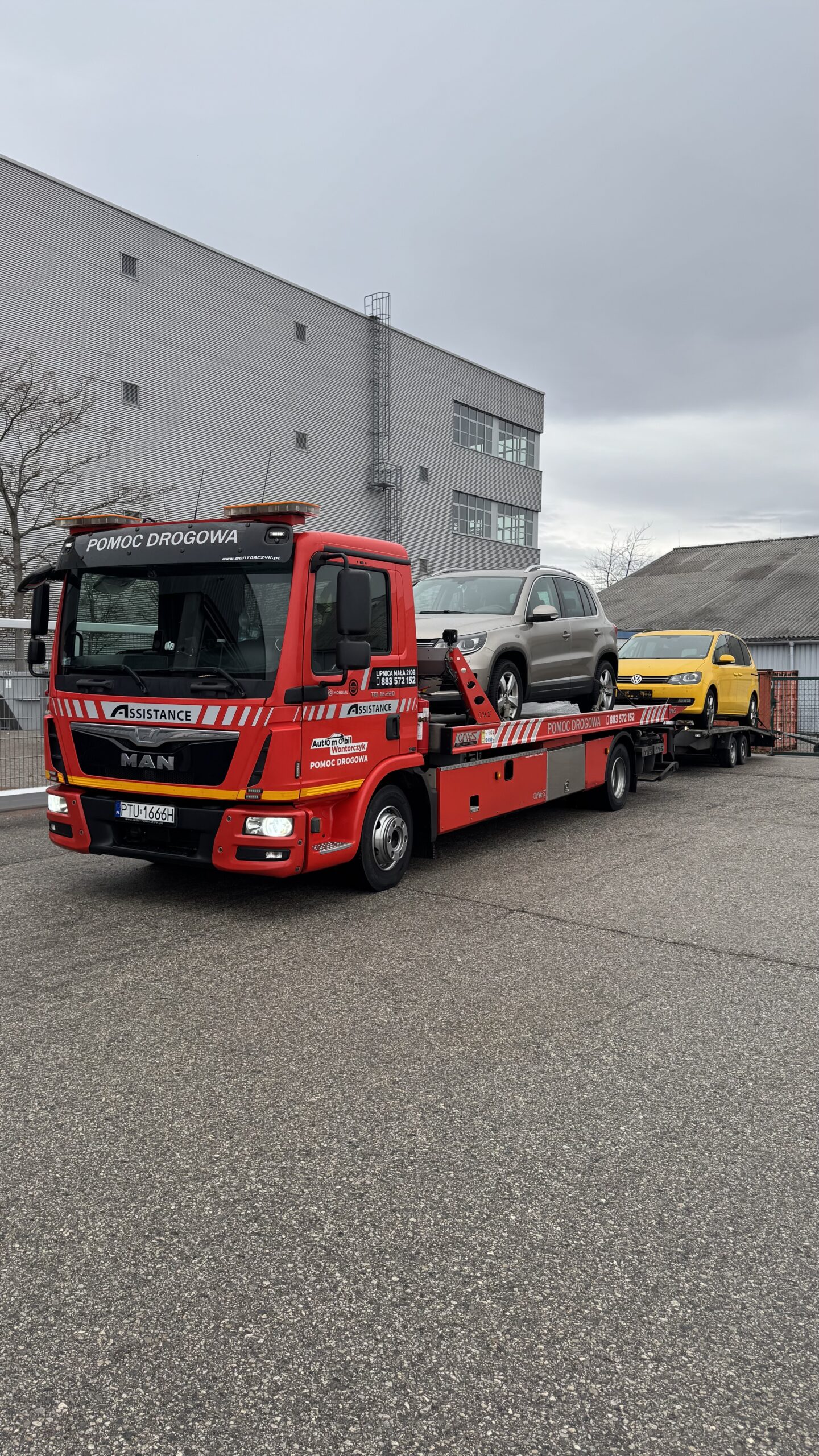 Czerwona laweta MAN pomocy drogowej przewożąca srebrny i żółty samochód na utwardzonym placu.