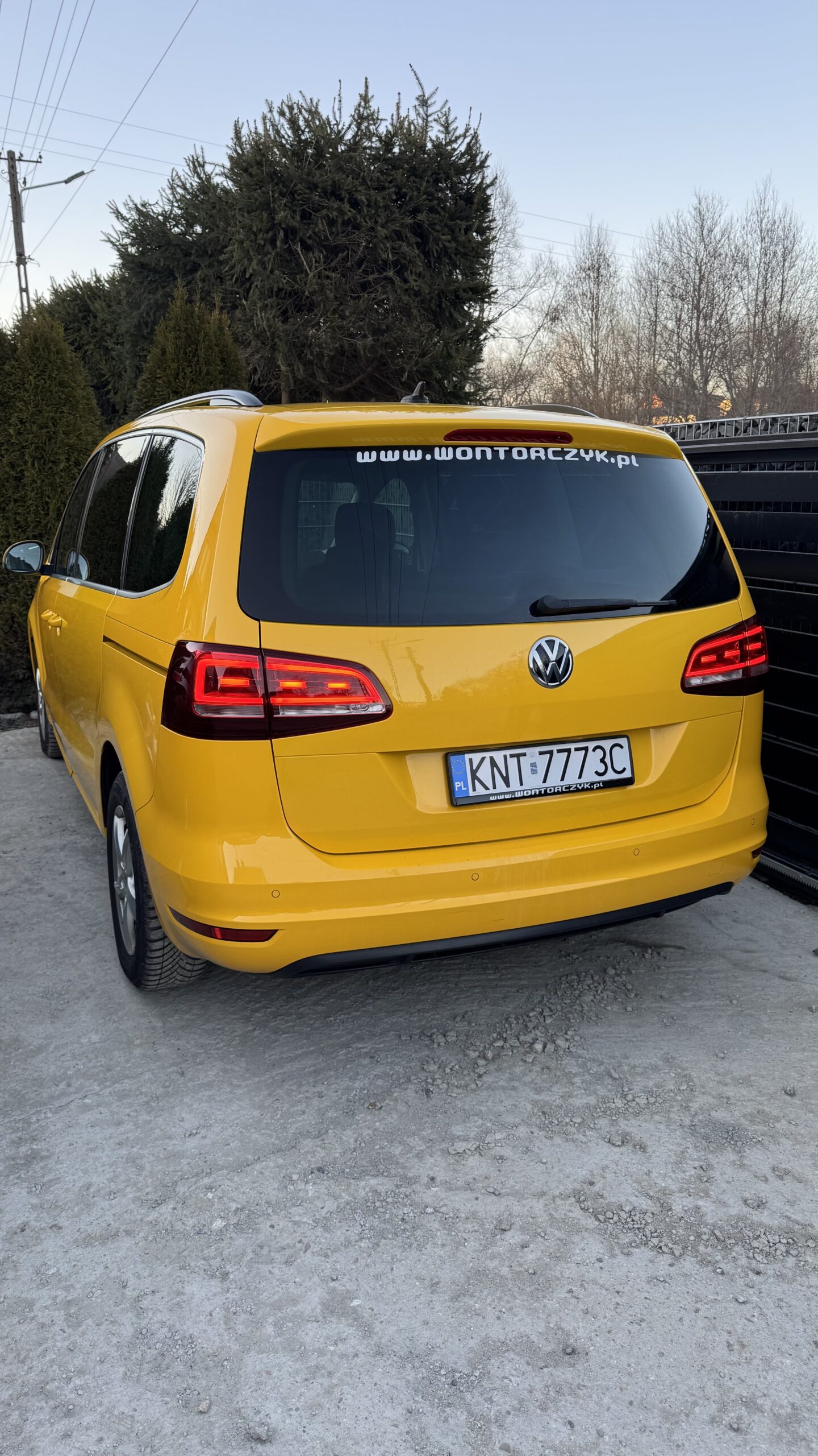 Żółty Volkswagen zaparkowany tyłem na żwirowym podjeździe, z widoczną naklejką firmową na tylnej szybie i rzędem drzew w tle.