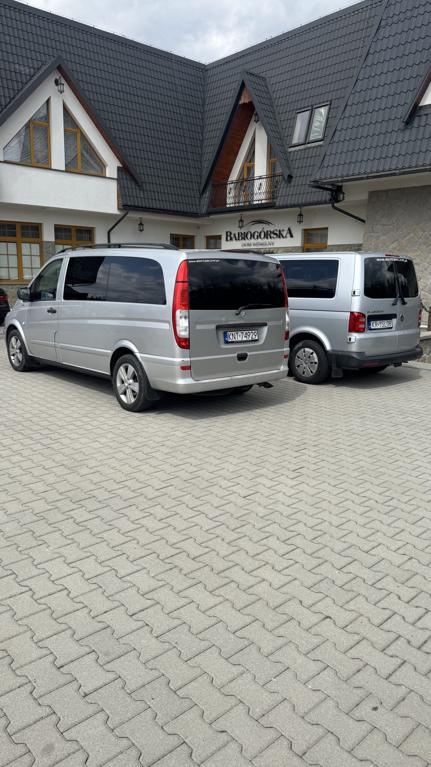Dwa srebrne busy zaparkowane tyłem na parkingu z kostki brukowej przed pensjonatem Babiogórska, z widocznym charakterystycznym dachem budynku w tle.