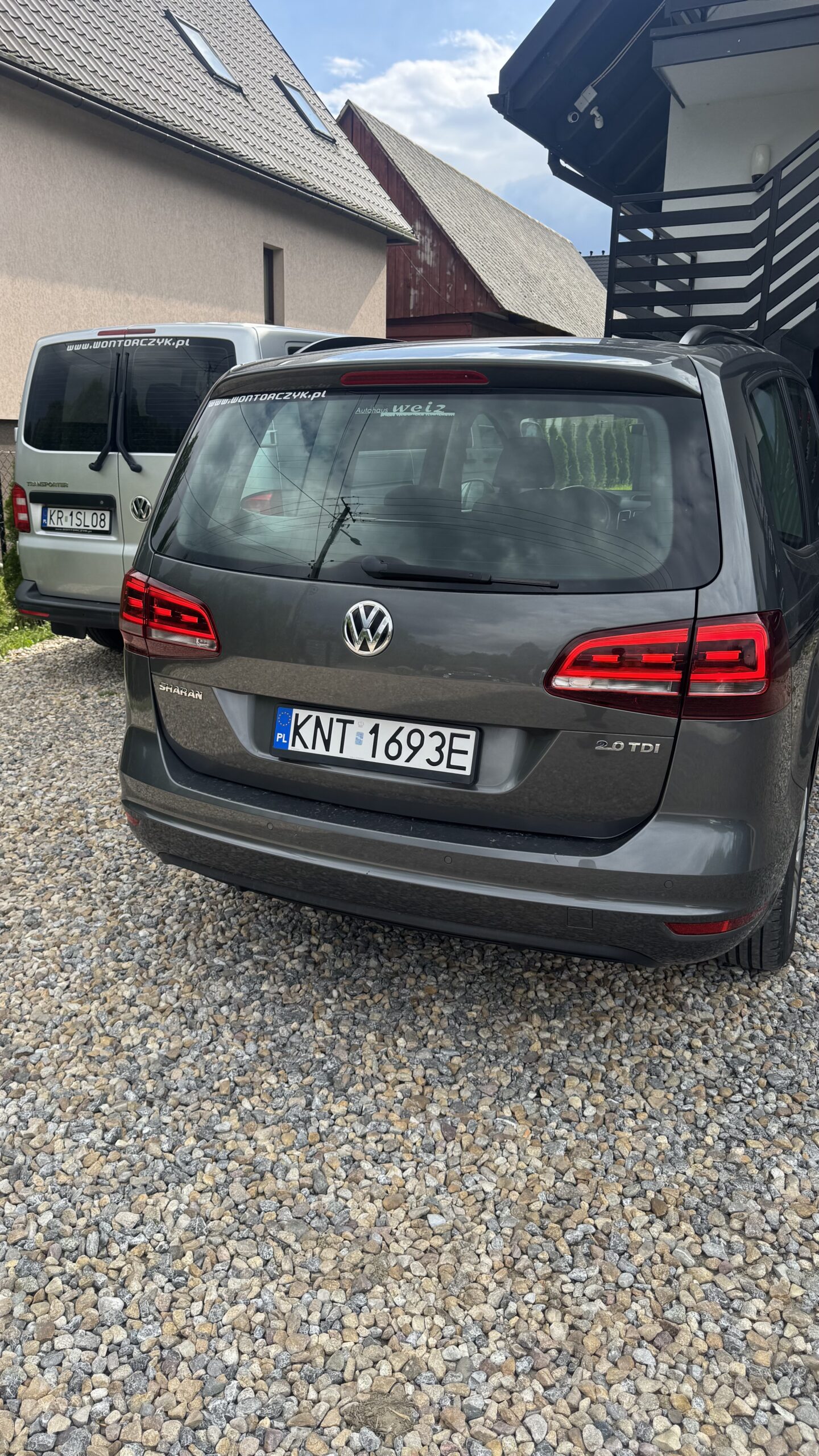 Szary Volkswagen Sharan zaparkowany tyłem na żwirowym podjeździe, obok stoi biały bus, a w tle widać dom i drewnianą stodołę.