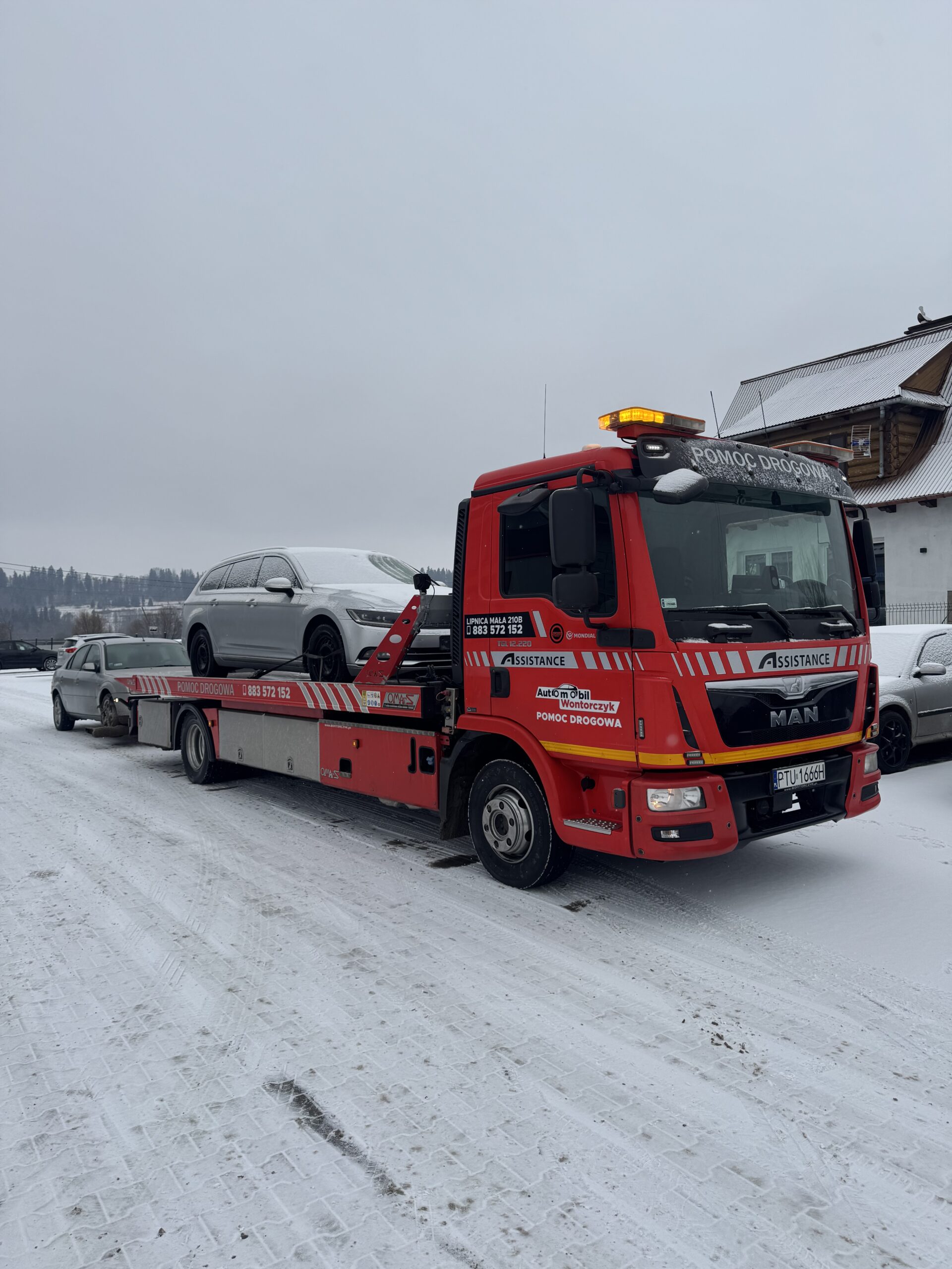Czerwona laweta MAN pomocy drogowej przewożąca srebrny samochód na zaśnieżonym terenie.