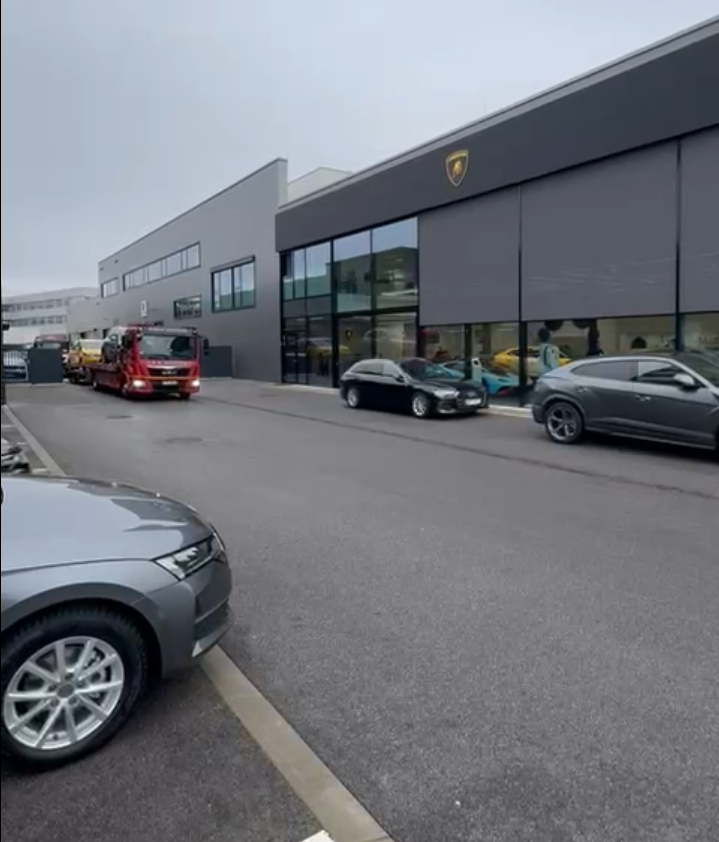 Czerwony holownik Automobil Wontorczyk jedzie ulicą obok nowoczesnego salonu Lamborghini z przeszkloną fasadą, przed którym stoją sportowe samochody.