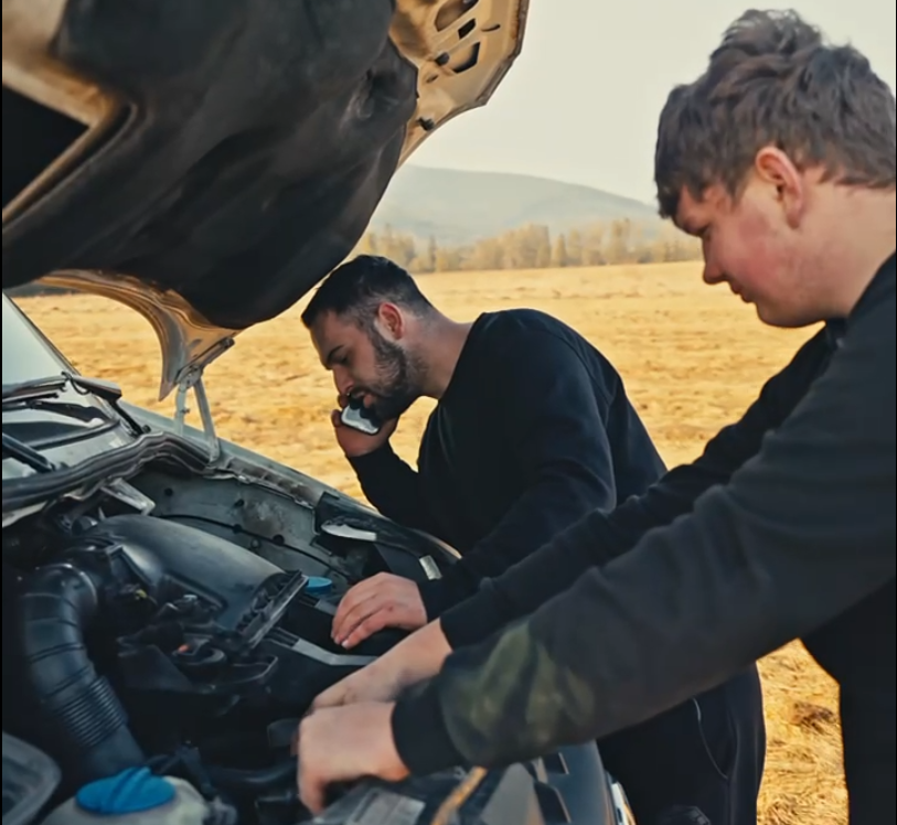 Dwóch mężczyzn pochylonych nad otwartą maską samochodu stojącego w polu; jeden sprawdza silnik, a drugi dzwoni po pomoc drogową.