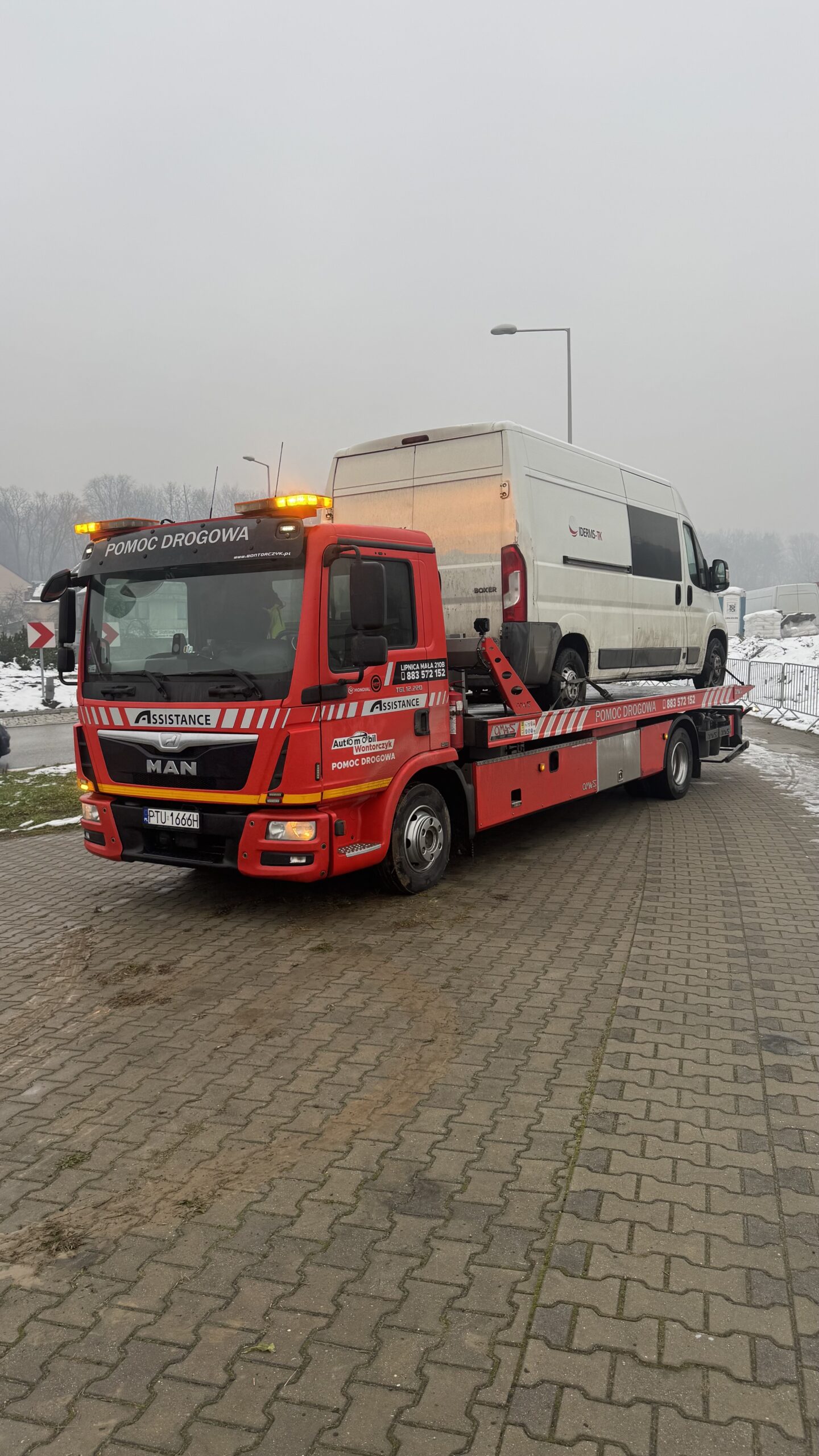 Czerwony holownik Automobil Wontorczyk przewozi na platformie biały bus dostawczy w zimowej, pochmurnej scenerii; pojazd stoi na kostce brukowej, a w tle widoczne są drzewa i śnieg.