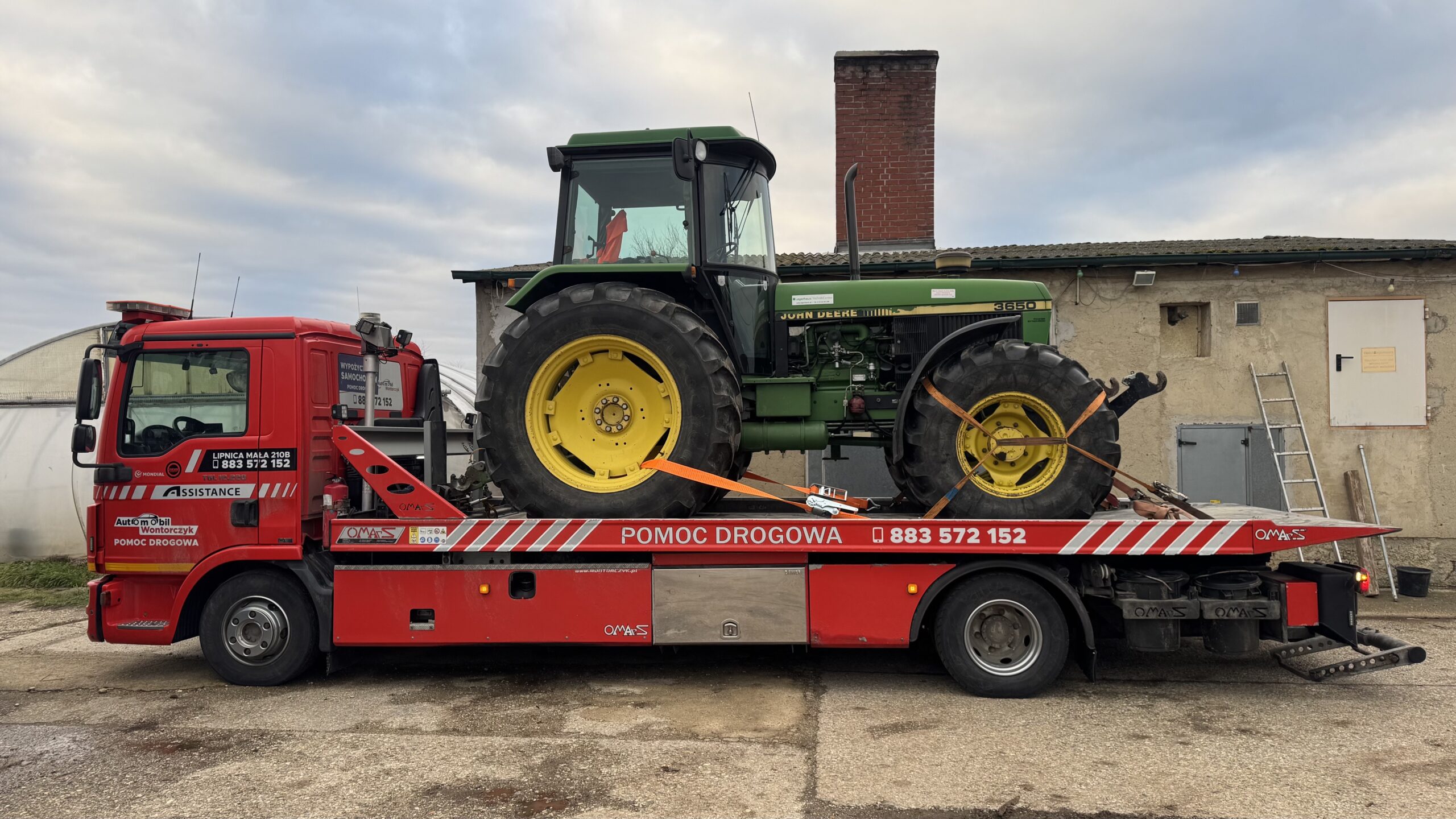 Czerwony holownik Automobil Wontorczyk przewozi na platformie duży zielony traktor John Deere, zabezpieczony pasami; pojazdy stoją na placu przy gospodarstwie z widocznym budynkiem i kominem w tle.