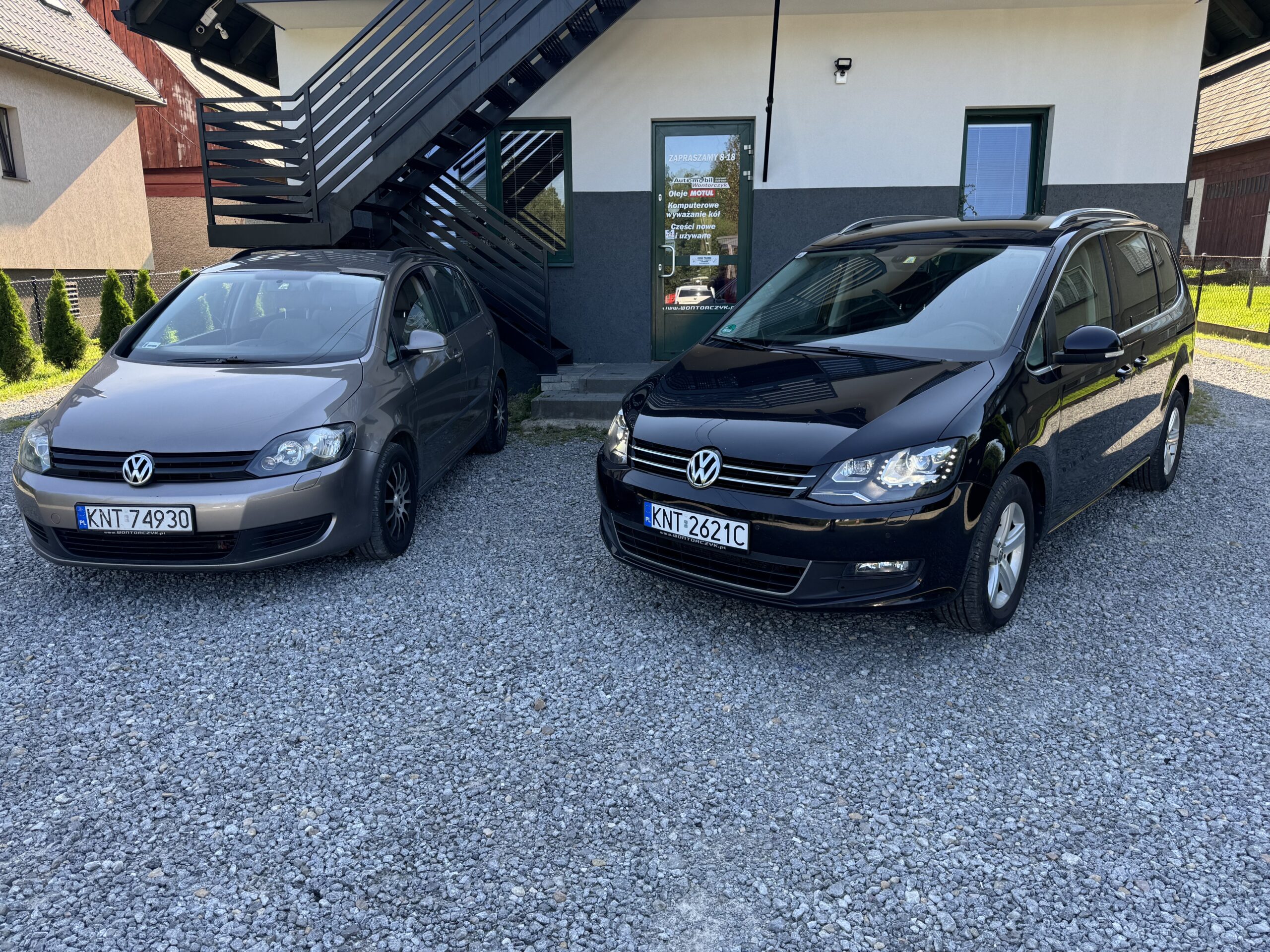 Dwa samochody marki Volkswagen – brązowy i czarny – zaparkowane na żwirowym placu przed wejściem do siedziby firmy Automobil Wontorczyk; w tle widać budynek z czarnymi schodami zewnętrznymi oraz drzwi prowadzące do biura.