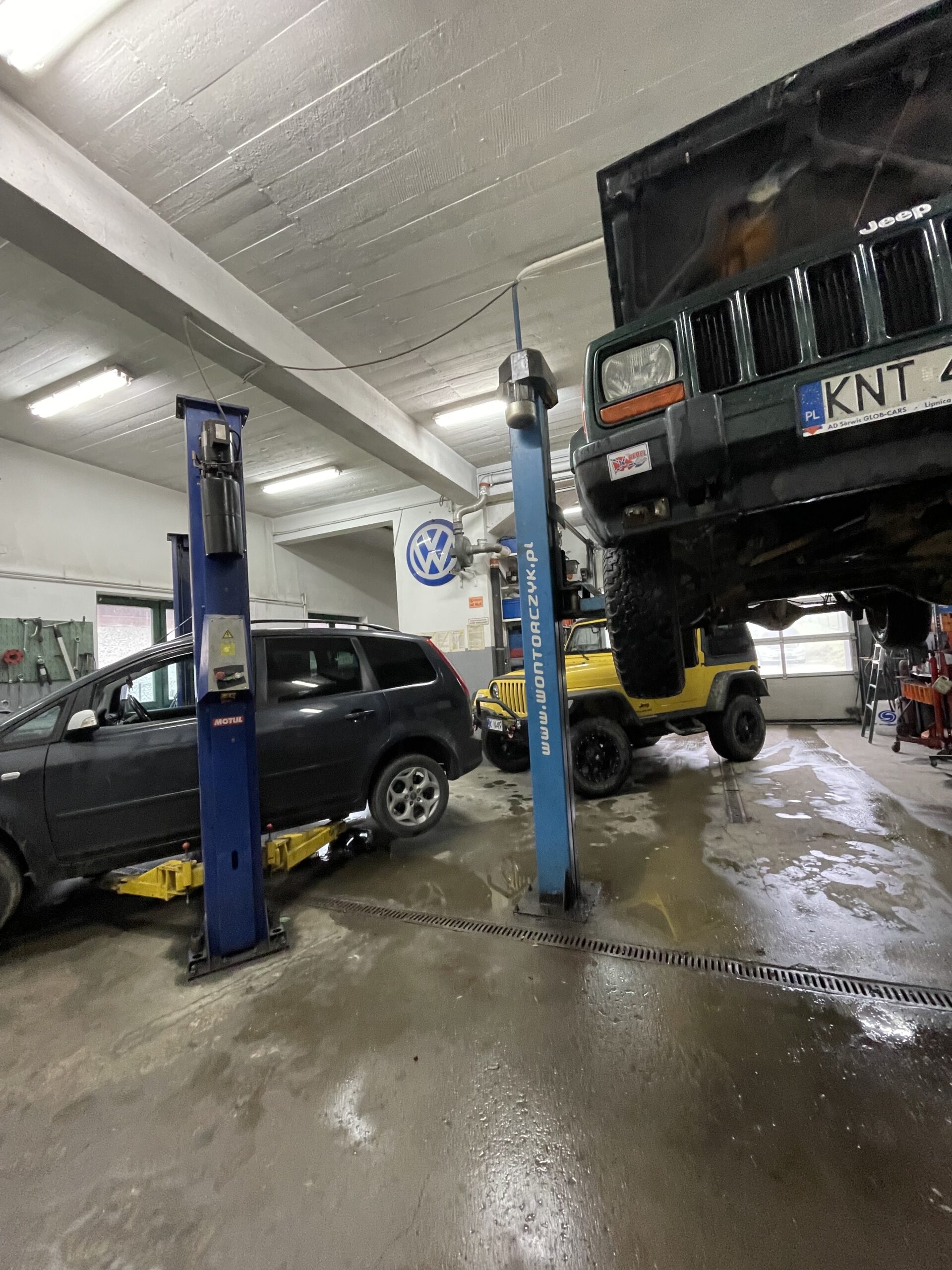 Wnętrze warsztatu samochodowego, w którym dwa auta stoją na podnośnikach — czarny samochód osobowy oraz zielony Jeep z otwartą maską — a obok nich znajduje się żółty samochód terenowy; podłoga jest mokra, a w tle widać narzędzia i wyposażenie serwisu.