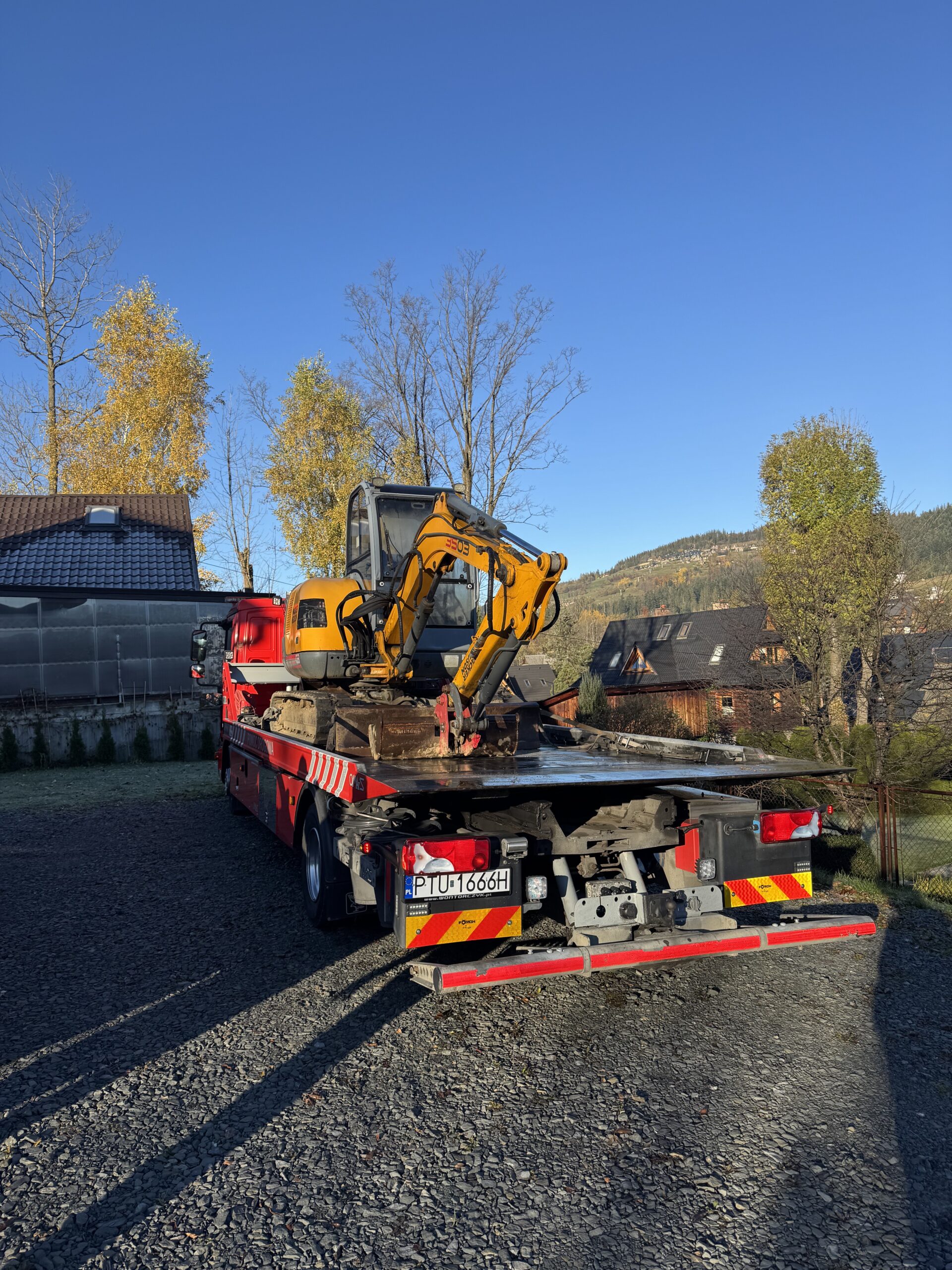 Czerwony holownik Automobil Wontorczyk przewozi na platformie żółtą minikoparkę; zdjęcie wykonane w słoneczny dzień, na tle domów i drzew o jesiennych liściach.