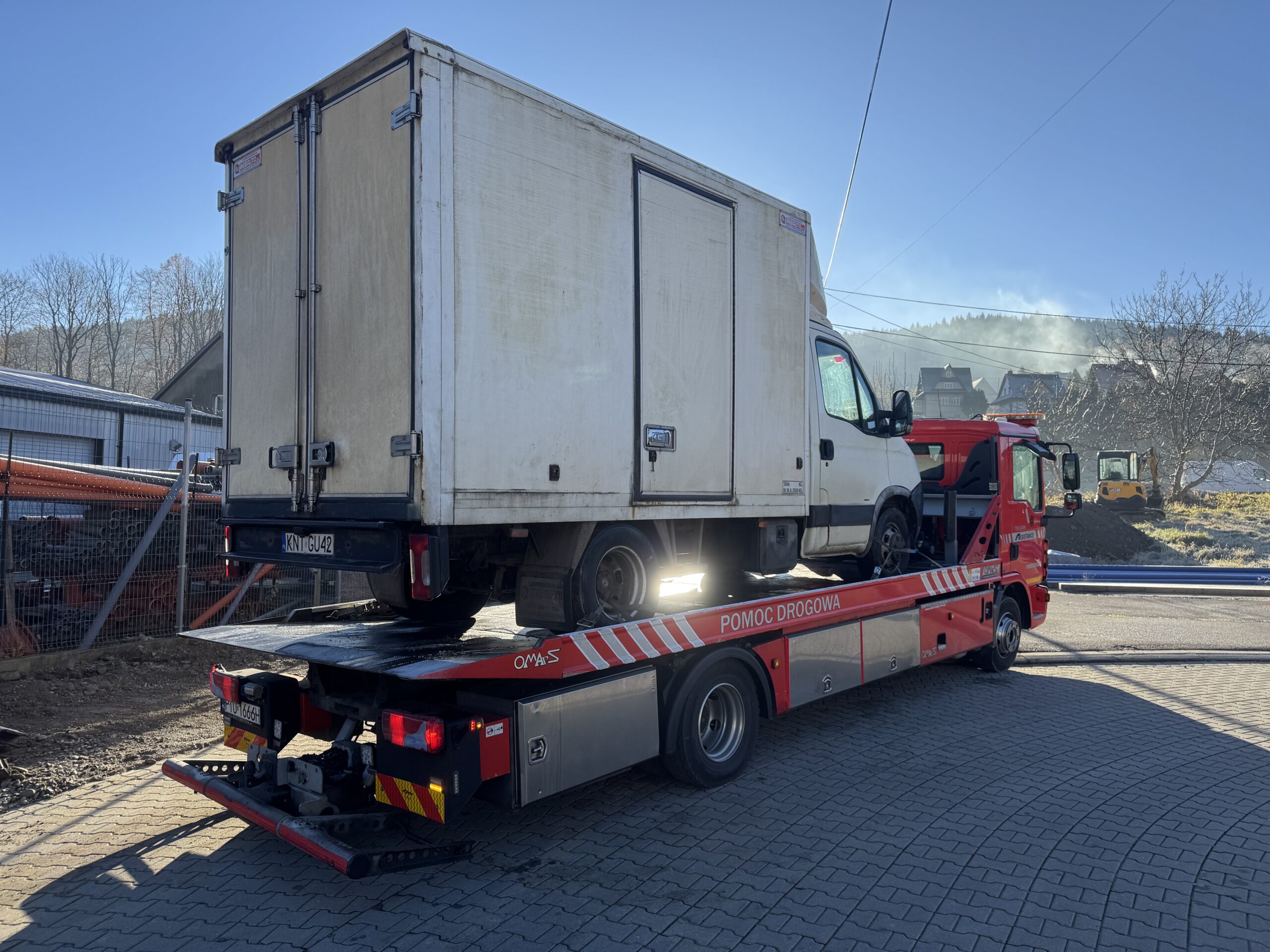 Czerwony holownik z napisem „Pomoc drogowa” przewozi na platformie białe auto dostawcze; pojazdy stoją na brukowanym placu, w tle widać zabudowania i wzgórza oświetlone zimowym słońcem.
