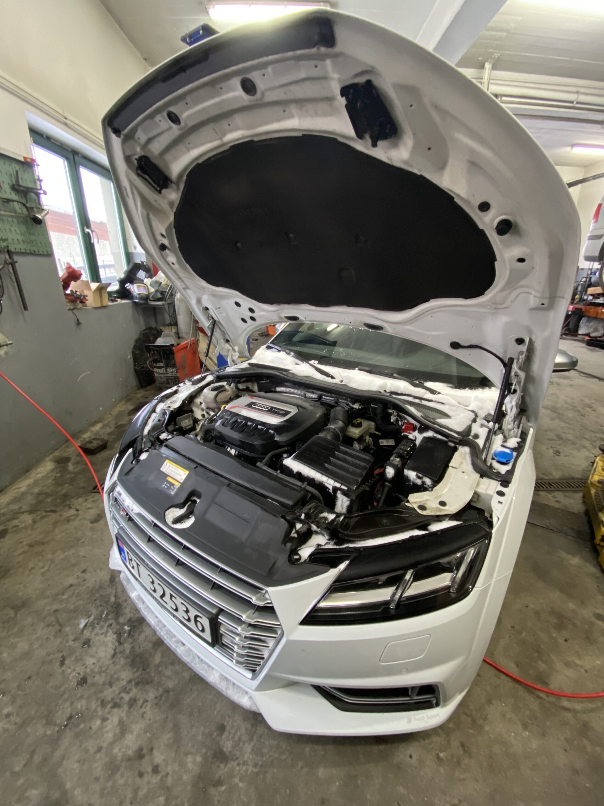 Biały samochód sportowy stoi w warsztacie z otwartą maską, odsłaniając silnik i elementy komory silnika, a wokół widoczne są narzędzia oraz wyposażenie serwisowe.