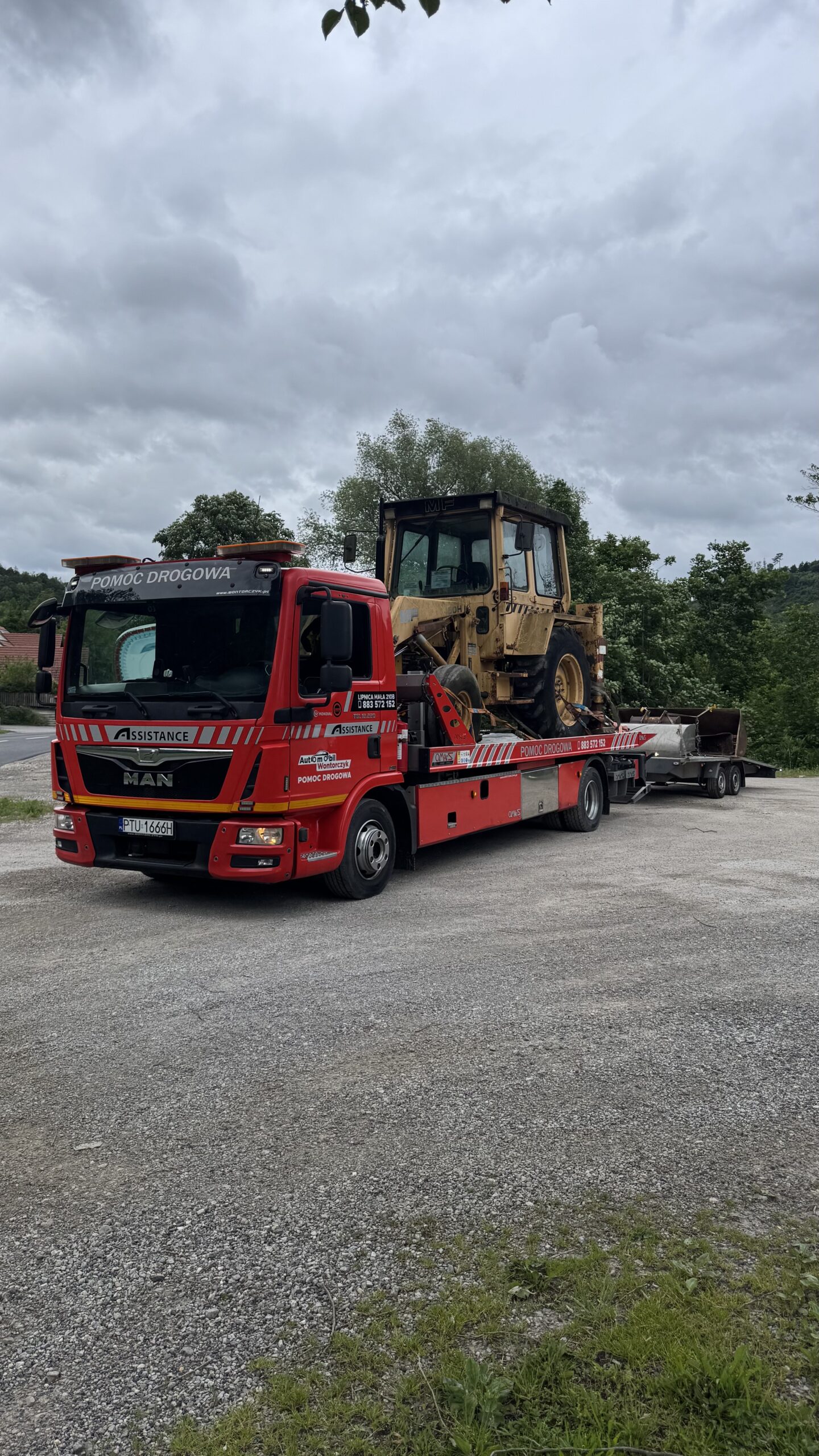 Czerwony holownik MAN z napisem „Pomoc drogowa” przewozi na platformie ciężką maszynę budowlaną; w tle widać drzewa i pochmurne niebo, a pojazd stoi na żwirowym placu.